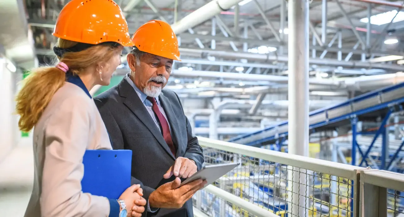 Two facility managers in hard hats reviewing a tablet on a walkway above a plant floor