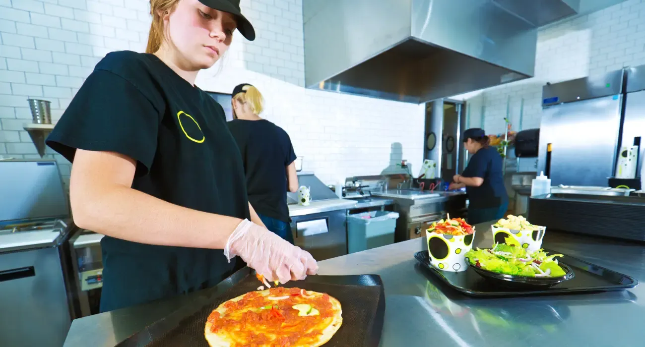 A kitchen worker preparing a pizza on a stainless steel counter in a commercial kitchen