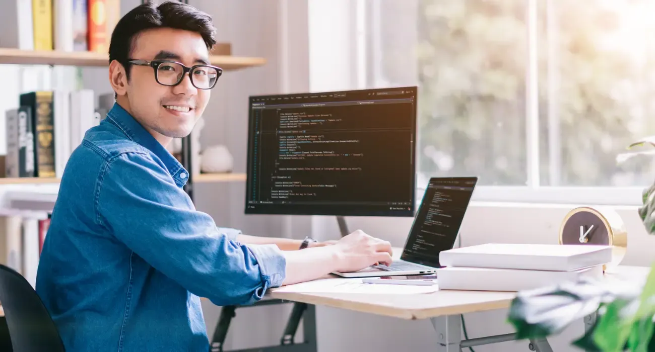 A software developer smiling at his desk in front of monitors showing code